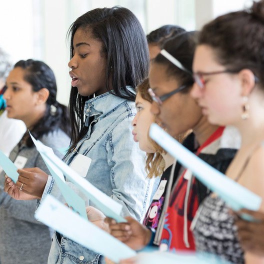 Group of young girls reading or singing from sheets of paper in an indoor setting.