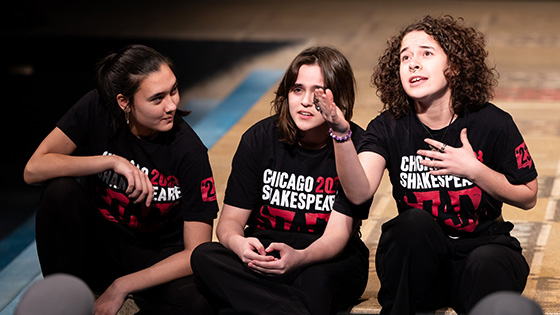 Three students wearing "Chicago Shakespeare SLAM" t-shirts sit on a stage floor, performing as if they are engaged in expressive conversation.