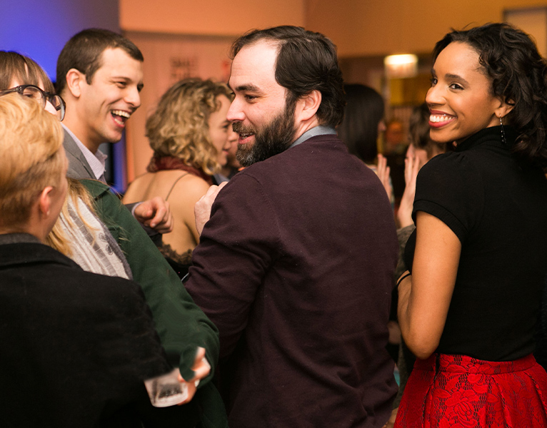 A group of people at a lively social gathering with several people interacting and smiling, prominently featuring a man with dark brown hair and beard and a woman with light brown skin and dark hair, both in profile view facing left.