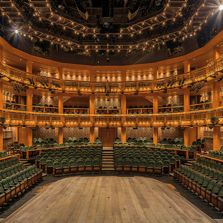 Theater interior with three levels of seating, green upholstered chairs, and wooden architectural details.