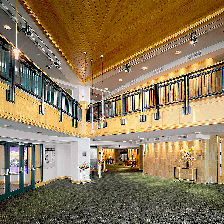 Spacious building interior lobby outside the Courtyard Theater, with a triangular wooden ceiling, a second-floor balcony with black railings, and dark green carpeted flooring. Glass double doors and floral arrangement are visible.