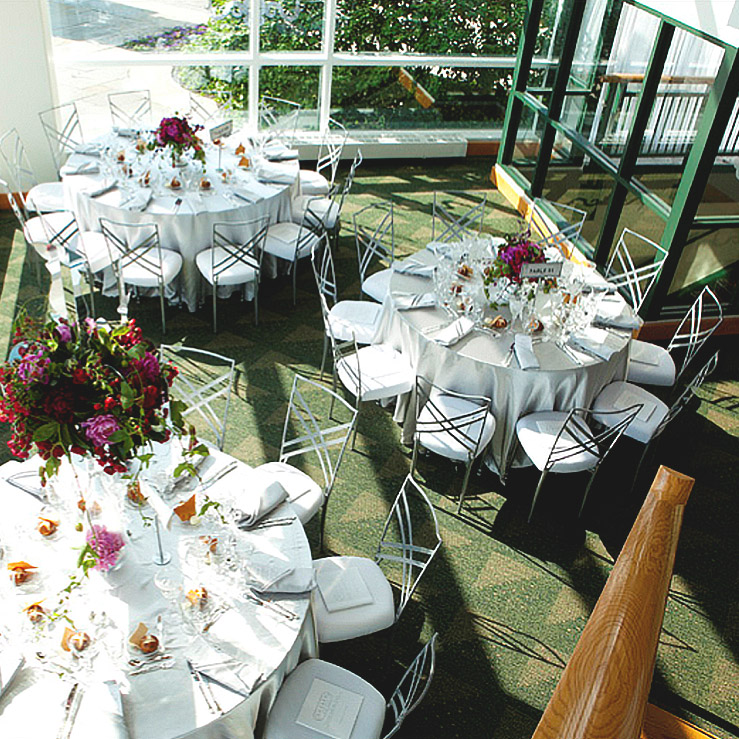 A formal indoor dining setup with round tables covered in white tablecloths, surrounded by metallic chairs with white cushions, and adorned with floral centerpieces and elegant place settings.