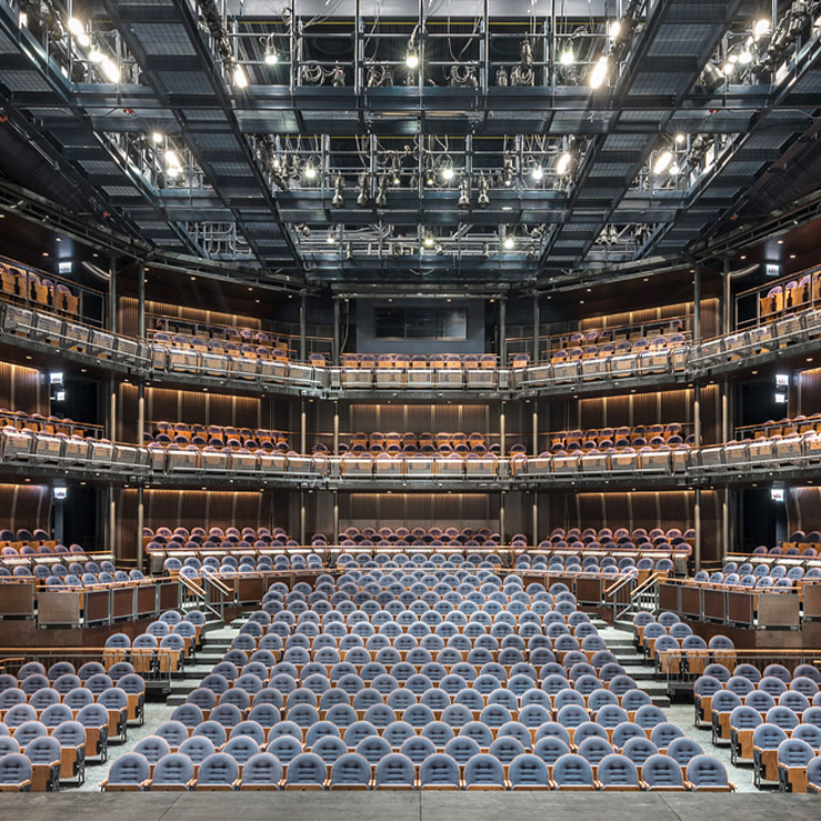Interior view of a large, modern multi-tiered theater from the stage, with blue cushioned chairs and warm lighting.