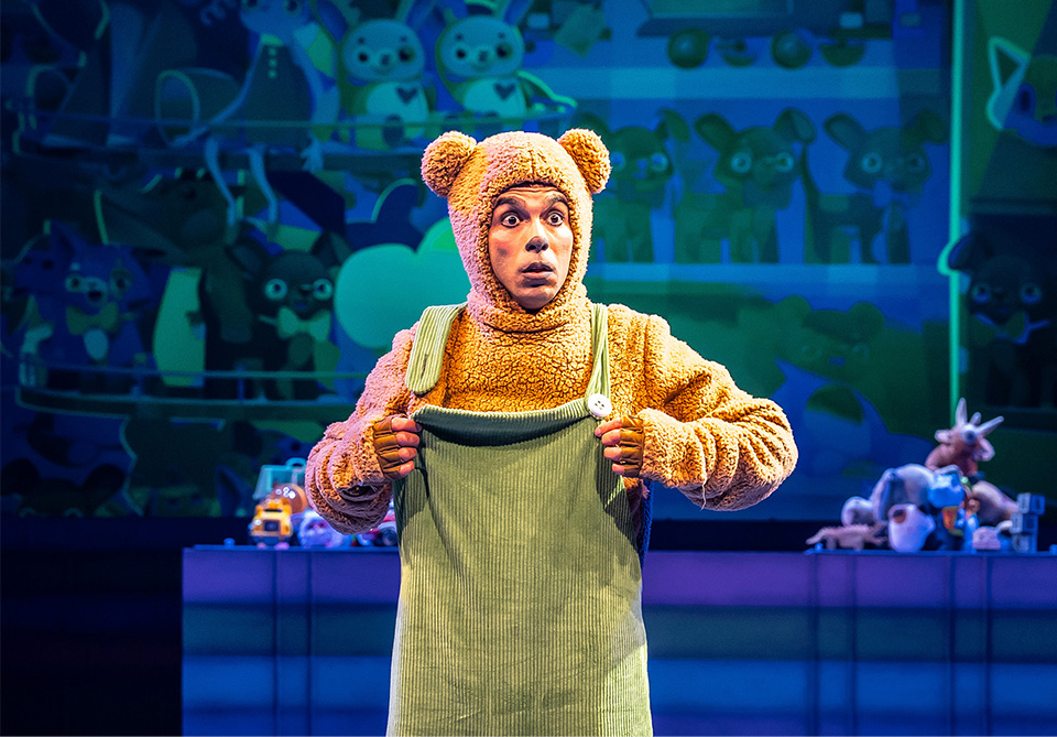 Person in a brown bear costume with green overalls performs on stage with a mural of cartoon animals in the background.