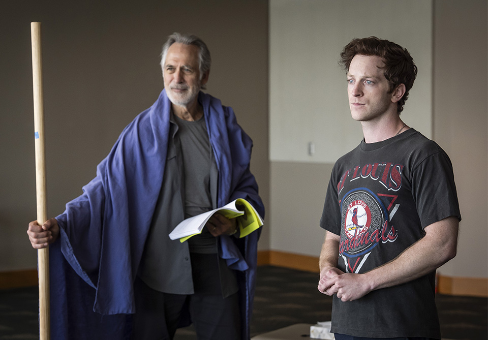 Two men standing indoors, one in a purple cloak holding a staff and book, and the other in a black St. Louis Cardinals T-shirt.