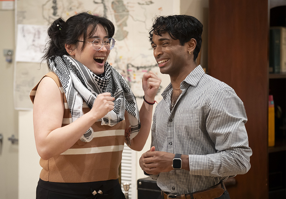 Two people smiling and interacting, one wearing glasses and a striped top with a scarf, the other in a checkered shirt.