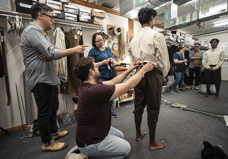 People in a costume fitting room, with one man adjusting another man's pants while others observe.