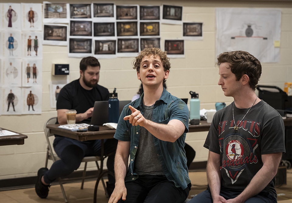 Three men in a creative workspace, one gesturing while talking, with design sketches on the wall behind them.