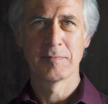 Close-up portrait of a person with gray hair wearing a dark maroon collared shirt against a dark background.