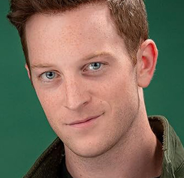Close-up of a person with short reddish brown hair, blue eyes, and a dark green jacket against a bright green background