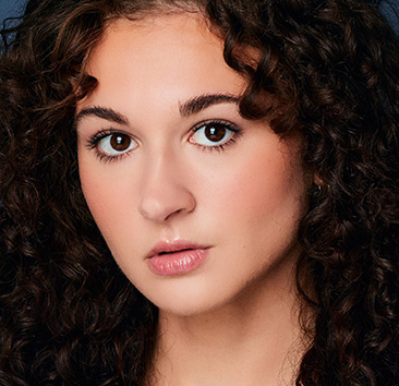 Close-up portrait of a person with curly dark brown hair, and dark brown eyes, looking directly at the camera