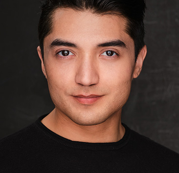 Portrait of a person with dark hair, wearing a black shirt, against a dark background