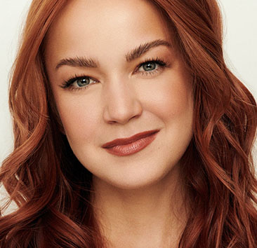 Close-up of a person with long, wavy red hair against an off-white background