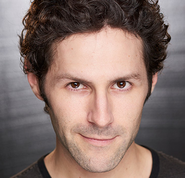 Close-up headshot of a person with short, dark, curly hair, brown eyes, light skin, and wearing a dark shirt, against a blurred dark gray background