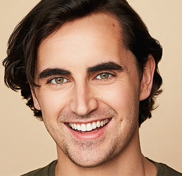 Close-up of a smiling man with dark brown hair and green eyes against a beige background