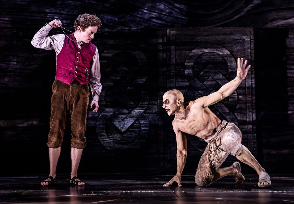 Frodo on left holding a chain, facing a crouching, shirtless Gollum on the right, on a dark stage with geometric patterns in the background.