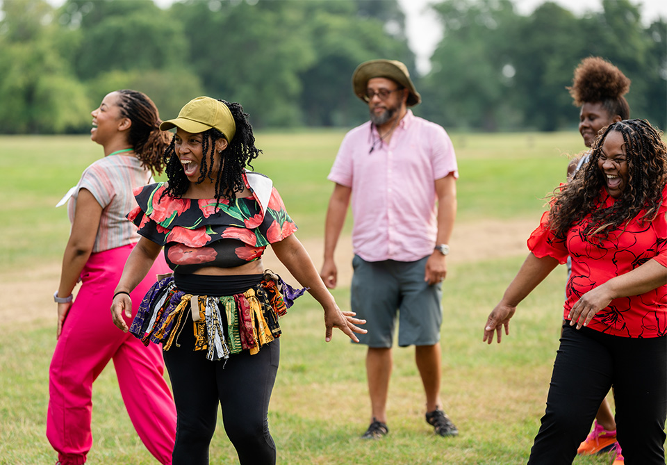 A group of five people joyfully dancing or engaging in an outdoor activity in a park, with three women in the foreground and a man and another woman in the background.