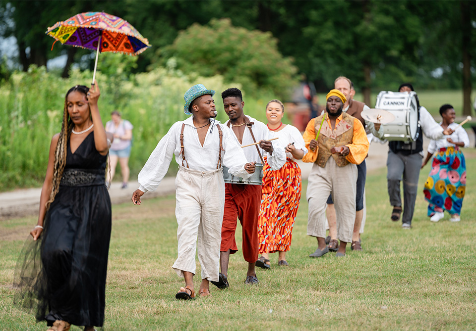 Parade of people in colorful attire walking on a grassy area, with greenery in the background.