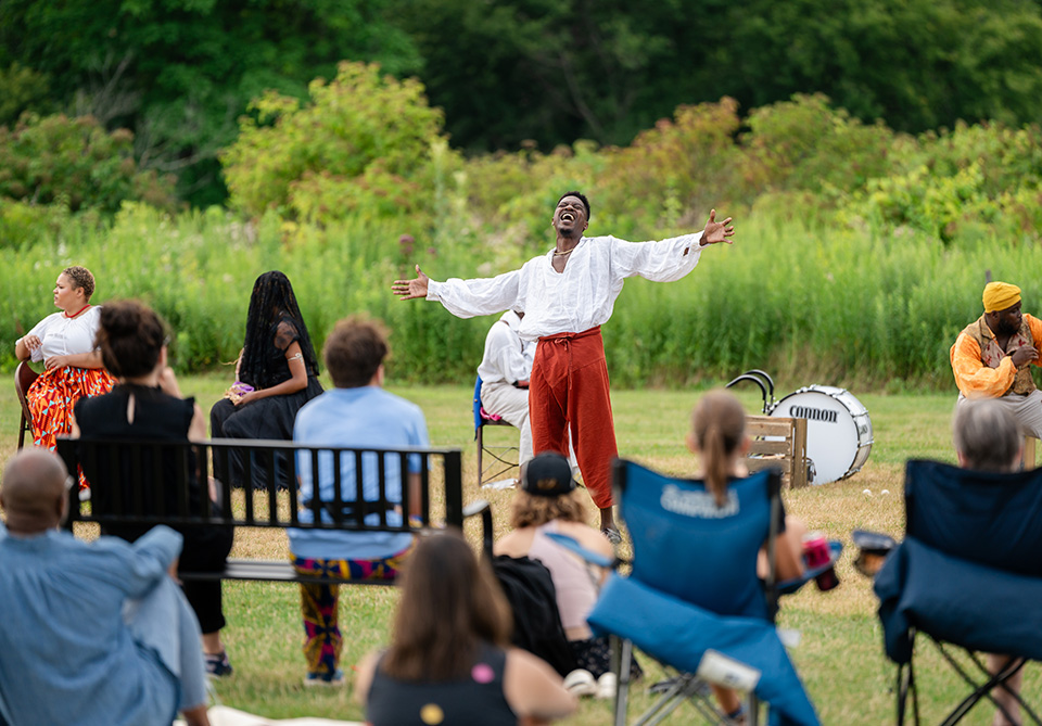 Outdoor theater performance with actors and audience; central actor stands with arms outstretched.