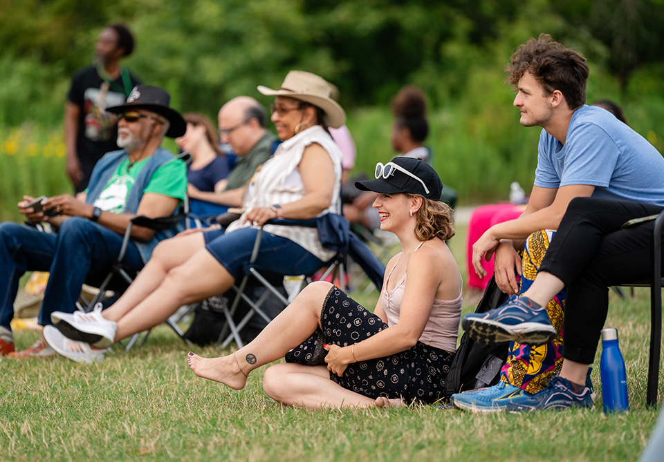 People seated on grass and chairs, watching an event in an outdoor park.