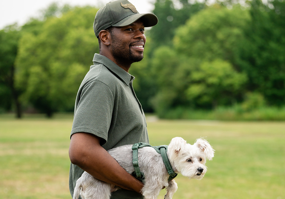 Man in a green polo shirt and cap holding a small grey dog in a park.