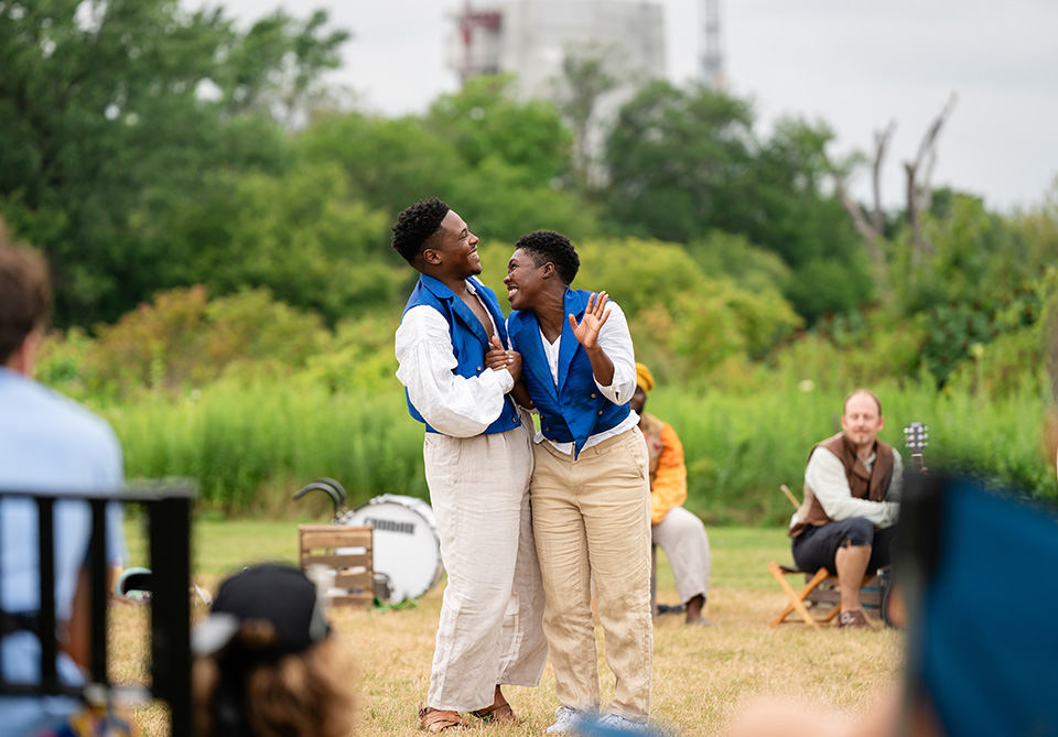 Two actors in blue vests and white pants embrace and smile outdoors, with others seated in the background.