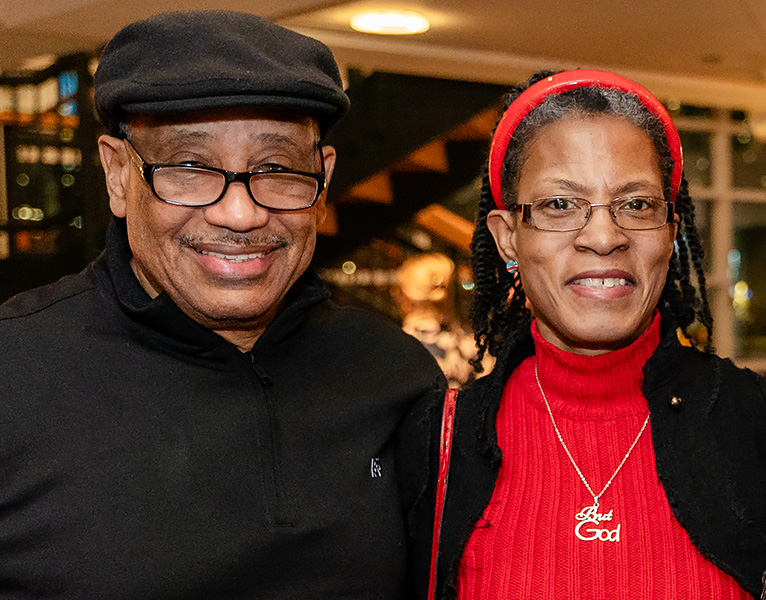 A couple smiles at camera, a man at left wearing a cap and glasses, and woman on right in a bright red shirt and headband.